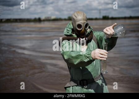 Un ambientalista maschile in una tuta protettiva verde e maschera a gas prende un campione di acqua in un lago inquinato. Rifiuti di produzione. Uno scienziato fa un Foto Stock