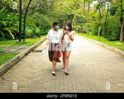 Medellin, Antioquia, Colombia - 6 gennaio 2021: Latin e le donne caucasiche che indossano una maschera facciale camminano in un parco con molti alberi Foto Stock