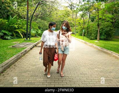 Medellin, Antioquia, Colombia - 6 gennaio 2021: Latin e le donne caucasiche che indossano una maschera facciale camminano in un parco con molti alberi Foto Stock