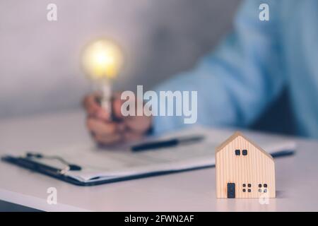 Agente immobiliare che tiene lampadina e accordo di contratto di proprietà immobiliare documento su scrivania, broker casa e investimento di pianificazione, ID pensiero uomo d'affari Foto Stock