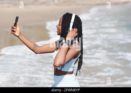 Sorridente giovane donna abbastanza in forma prendendo selfie sulla spiaggia dopo jogging al mattino Foto Stock