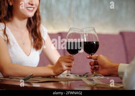 Immagine ravvicinata di una giovane coppia che brinda con bicchieri di rosso vino seduto al tavolo del ristorante Foto Stock