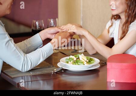 Immagine ritagliata di giovane donna che tiene le mani di fidanzata e. dicendole i complimenti e le parole di amore quando sono seduto al tavolo del ristorante Foto Stock