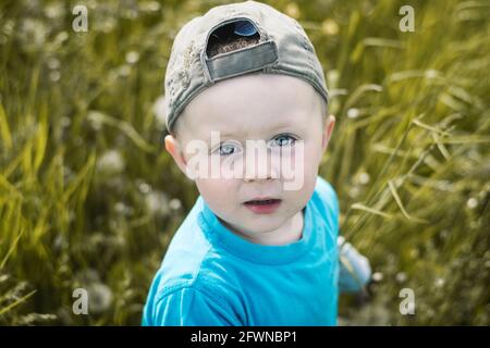 Il ragazzo caucasico di due anni con berretto da baseball e t-shirt blu guarda alla fotocamera. Sullo sfondo si difocusava l'erba con colori smorzati. Foto Stock