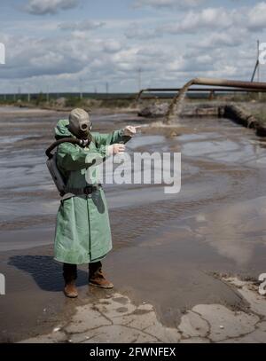 Un ambientalista maschile in una tuta protettiva verde e maschera a gas prende un campione di acqua. Lo scienziato sta facendo uno studio tossicologico. Post apocalisse Foto Stock