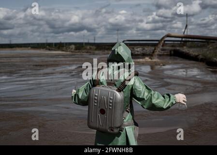 Un ambientalista maschile in una tuta protettiva verde e maschera a gas prende un campione di acqua in un lago inquinato. Rifiuti di produzione. Uno scienziato fa un Foto Stock