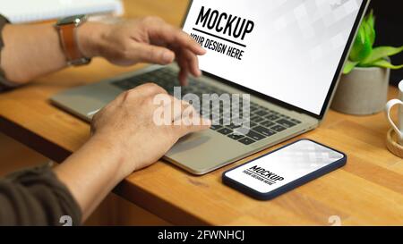 Ritaglio di mani maschili digitando sulla tastiera del computer portatile su un tavolo di legno con smartphone, ritaglio percorso Foto Stock