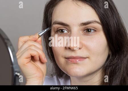 Una donna bruna le ha strappato le sopracciglia davanti ad uno specchio. Correzione della forma delle sopracciglia con pinzette da sole Foto Stock