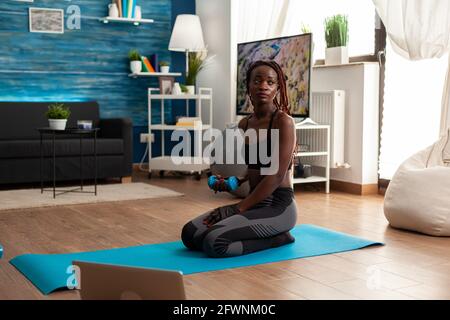 Fit atletica donna nera attiva flettere le braccia lavorando bicipite, seduta su tappetino yoga in casa soggiorno esercizio seguendo le istruzioni video dal computer portatile, avendo uno stile di vita sano. Foto Stock