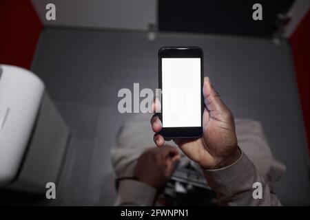 Vista dall'alto di un uomo afroamericano irriconoscibile che usa lo smartphone mentre si siede sulla toilette in toilette pubblica, spazio per fotocopie Foto Stock