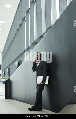Ritratto grafico grandangolare di sicuro uomo d'affari afro-americano che parla smartphone in piedi su sfondo nero nella sala dell'ufficio Foto Stock
