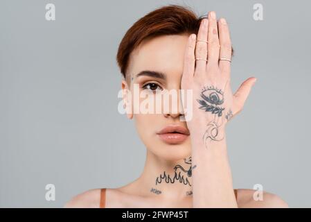 giovane donna tatuata che guarda la macchina fotografica e copre il viso con isolato a mano su grigio Foto Stock