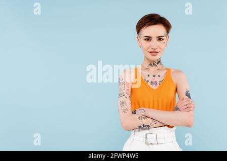 donna contenta e tatuata con canotta in maglia arancione in piedi con bracci incrociati isolati in blu Foto Stock