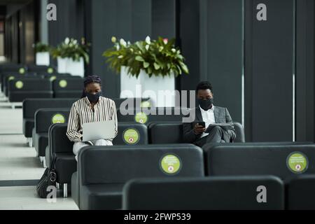 Ritratto grafico grandangolare di uomo d'affari afroamericano indossando maschera mentre lavorare con il computer portatile in sala d'attesa in aeroporto con social distanziamento Foto Stock