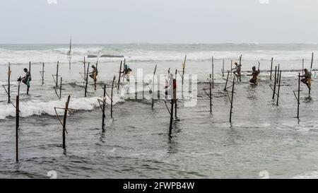 Mirissa, Sri Lanka - 07 26 2020: Gruppo di pescatori tradizionali di stendere che tengono canne da pesca seduti su un bastone croci sopra il patie di superficie dell'acqua di mare Foto Stock