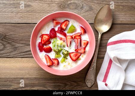 Porridge di farinata d'avena con fette di kiwi, fragole in ciotola rosa, cucchiaio, tovagliolo con strisce rosse su sfondo di legno Vista dall'alto piatto piatto piatto cibo vegetariano Foto Stock