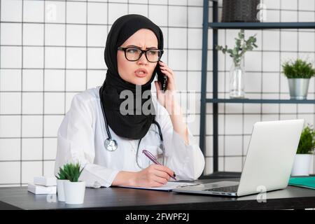 Femmina scienziato velata seduta sulla sua scrivania guardando laptop apre la sua bocca con turbato viso emozione durante la conversazione su un telefono cellulare Foto Stock