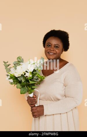 sorridente afroamericano di mezza età donna in abiti pastello con bouquet di fiori isolati su beige Foto Stock