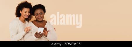 figlia adulta afroamericana che punta con il dito allo smartphone nelle mani di madre di mezza età su sfondo beige, banner Foto Stock