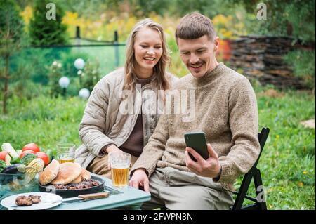 Una giovane coppia ha cenato con la famiglia lontano dalla città. Picnic con carne alla griglia e birra. Scattano foto di natura bellissima per i loro social media Foto Stock