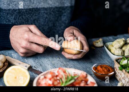 un giovane caucasico taglia un panino per preparare un panino o degli antipasti vegani, seduti ad un tavolo di legno grigio, accanto ad una ciotola con un sa di pomodoro Foto Stock