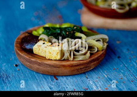 closeup di un piatto di legno, su un tavolo di legno blu, con alcuni dadi di tofu, tagliatelle di farro, fagioli di edamame e kale, e un altro piatto vegano nel backgr Foto Stock