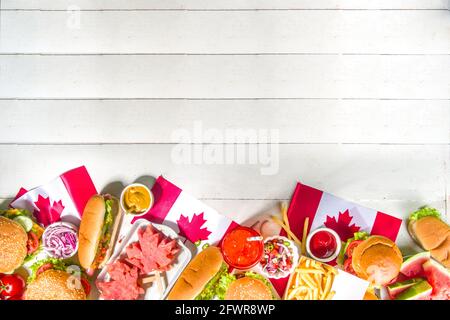 Set di vari piatti barbecue del Canada Day. Tavolo da picnic con cocomero a foglia d'acero, bandiere, hamburger, hot dog, patatine fritte e salse, bevande fredde, whi Foto Stock