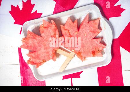 Canada Day Concept, tradizionale foglia d'acero fatta da fette di anguria su piccolo vassoio, su bandiere canadesi e tavolo bianco in legno vista dall'alto Foto Stock
