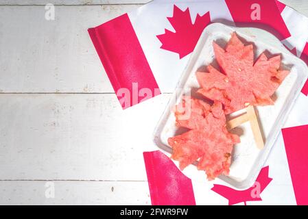 Canada Day Concept, tradizionale foglia d'acero fatta da fette di anguria su piccolo vassoio, su bandiere canadesi e tavolo bianco in legno vista dall'alto Foto Stock