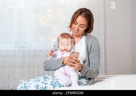 Una madre tiene un bambino addormentato tra le braccia e gli mostra qualcosa sul suo smartphone. Primo piano verticale. Interni domestici. Foto Stock