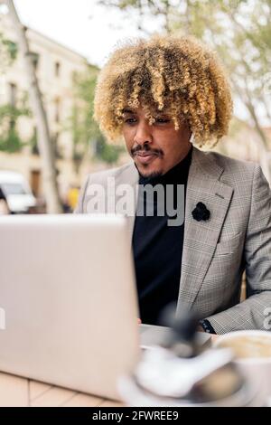 Uomo d'affari africano con capelli afro seduto in caffetteria e utilizzando il suo laptop. Indossa gli auricolari e ascolta la musica. Foto Stock