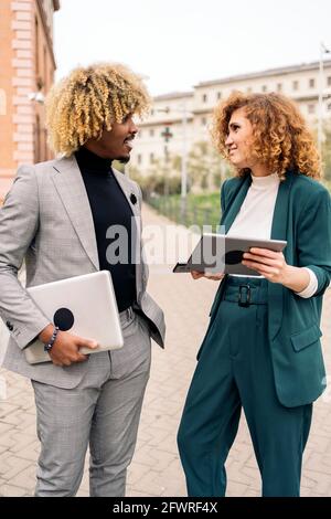 Felice uomo d'affari afro che indossa abiti formali parlando con il suo collega e tenendo un computer portatile in strada. Foto Stock