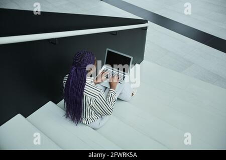 Vista posteriore grafica di una donna d'affari afro-americana con un computer portatile mentre si siede sulle scale nella sala d'ufficio, spazio per la copia Foto Stock