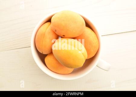 Diverse albicocche di ananas gialle mature in una tazza di ceramica bianca, primo piano, su un tavolo di legno bianco, vista dall'alto. Foto Stock