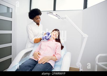 Likable giovane donna africana cosmetologist beautician facendo il trattamento facciale con spatola ultrasonica a giovane donna caucasica. Pulizia della pelle del viso e trattamento di pulizia con dispositivo a ultrasuoni Foto Stock