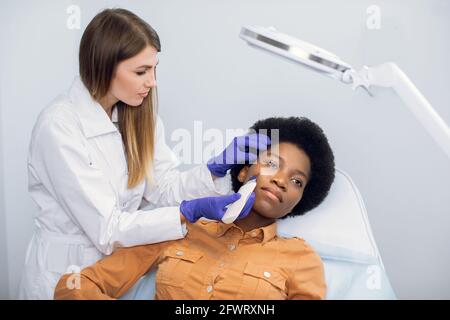 Concetto di bellezza e cura del viso. Dottore femminile cosmetologist in guanti blu rende ecografia procedura di pulizia della pelle del viso di giovane donna piuttosto africana in salone di bellezza Foto Stock