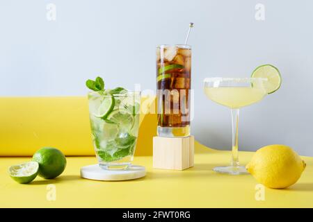 Mojito classico, cuba libre, cocktail margarita con lime e limone su sfondo giallo di colore moderno. Tre bevande fresche estive per le feste Foto Stock