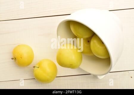 Numerose prugne gialle mature con una tazza bianca, primo piano, su un tavolo di legno bianco, vista dall'alto. Foto Stock