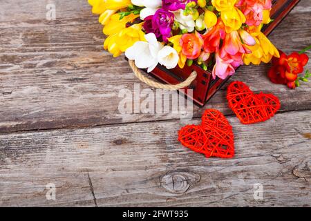 Freesia multicolore in una scatola e due cuori su un vecchio sfondo di legno. Bellissimi fiori. Foto Stock
