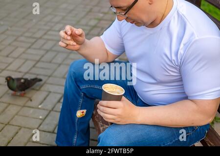 un ragazzo con bicchieri, tenendo una tazza di caffè e lasciando cadere un pezzo di cibo, un panino, una ciambella dalla mano a terra durante il pranzo, seduto su una panchina Foto Stock