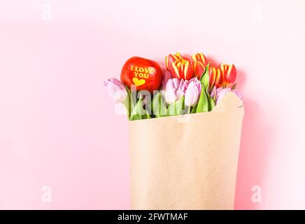 Bouquet di bellissimi fiori di tulipano primavera avvolti in borsa di carta artigianale e mela rossa con parole ti amo su morbido sfondo rosa. Foto Stock