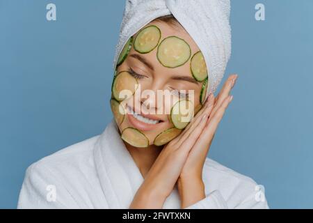 La donna soddisfatta inclina la testa sui palmi premuti insieme tiene gli occhi chiudersi e sorrisi delicatamente fa la maschera facciale applica le fette di cetriolo sulla faccia gode Foto Stock
