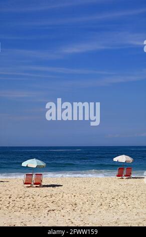 Paia di sedie da spiaggia rosse vuote con ombrelloni sul spiaggia di sabbia di fronte alle onde che si sprigionano sul vivido oceano blu Foto Stock