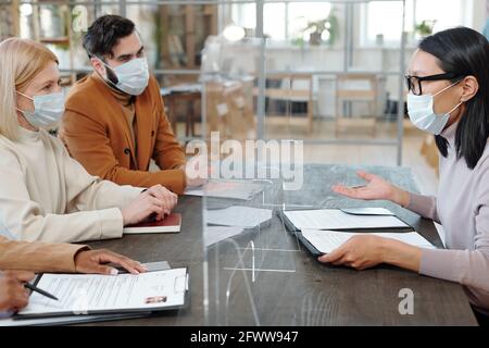 Perplessa giovane donna asiatica in maschera seduta dietro lo schermo di barriera e negoziando il contratto con i recruiters al colloquio di lavoro Foto Stock
