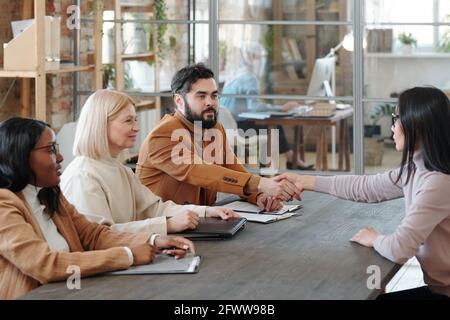 Recruiter bearded scuotendo la mano della donna asiatica mentre si congratula con lei con un colloquio di lavoro riuscito in ufficio Foto Stock