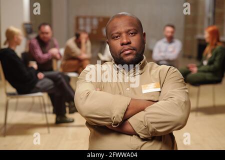 Uomo africano serio che attraversa le braccia per petto contro il gruppo di persone che parlano di problemi di trir Foto Stock