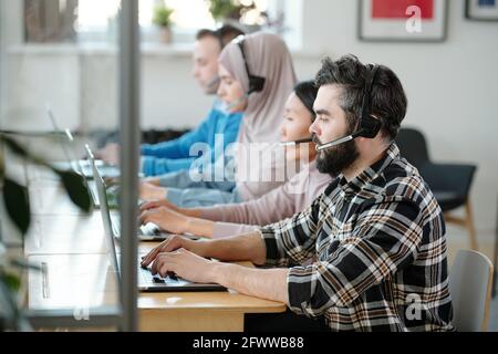 Fila di operatori di call center multietnici impegnati in cuffia con microfono che lavora con le applicazioni del cliente Foto Stock