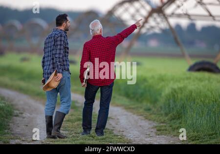 Two men walking next to field looking pointing showing center pivot irrigation system. Farmers surveying land modern agriculture. Foto Stock