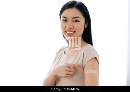 Felice bella giovane donna asiatica, vestita in t-shirt, con un gesso adesivo sulla spalla dopo essere stato vaccinato di COVID 19 o influenza, guardando la macchina fotografica, sorride volentieri Foto Stock