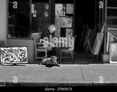 Un artista anziano lavora su un disegno mentre si siede all'ombra davanti ad una galleria d'arte con il suo cane da compagnia al suo fianco a Santa Fe, New Mexico. Foto Stock